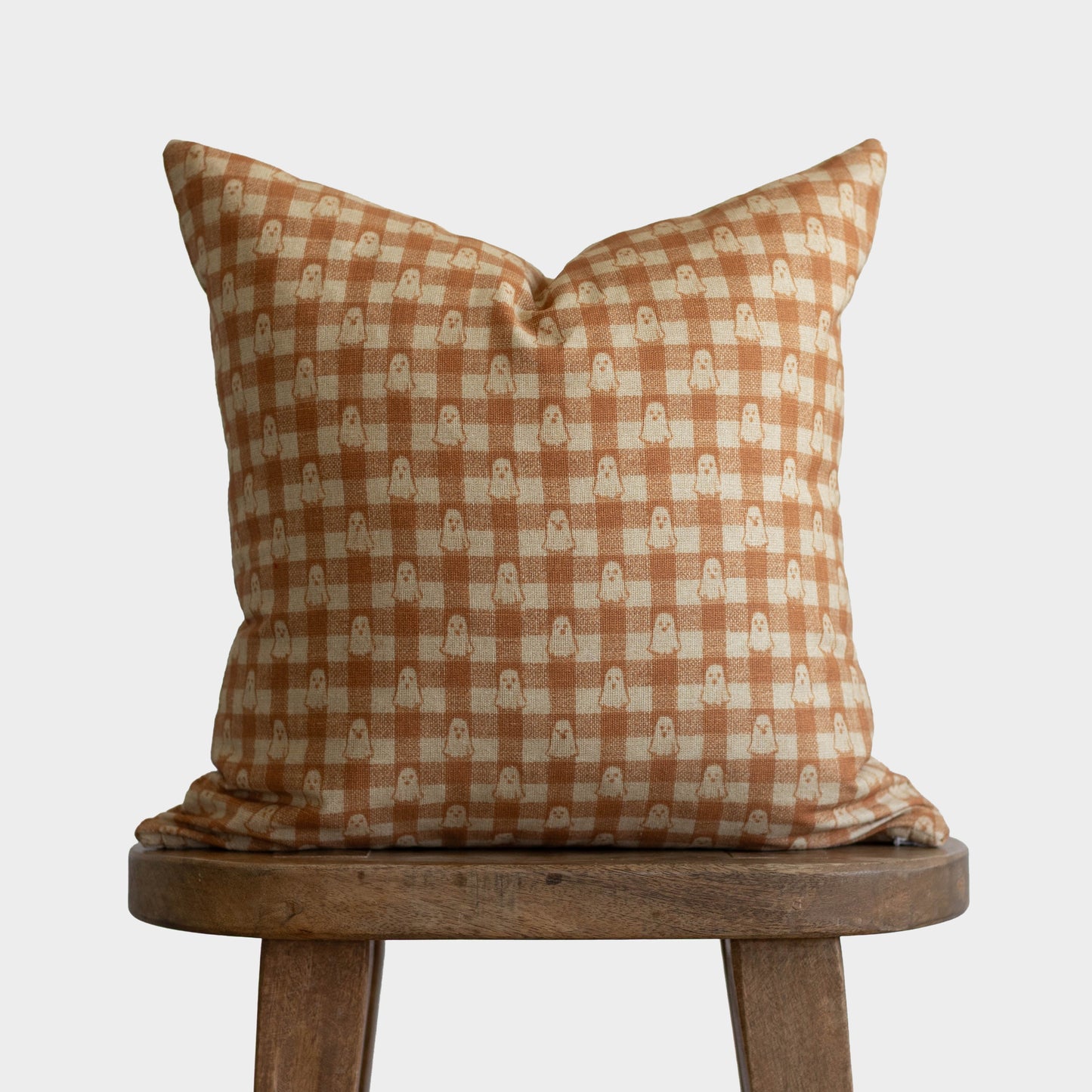 Rust and cream gingham pillow with small ghost motifs, displayed on wooden stool.