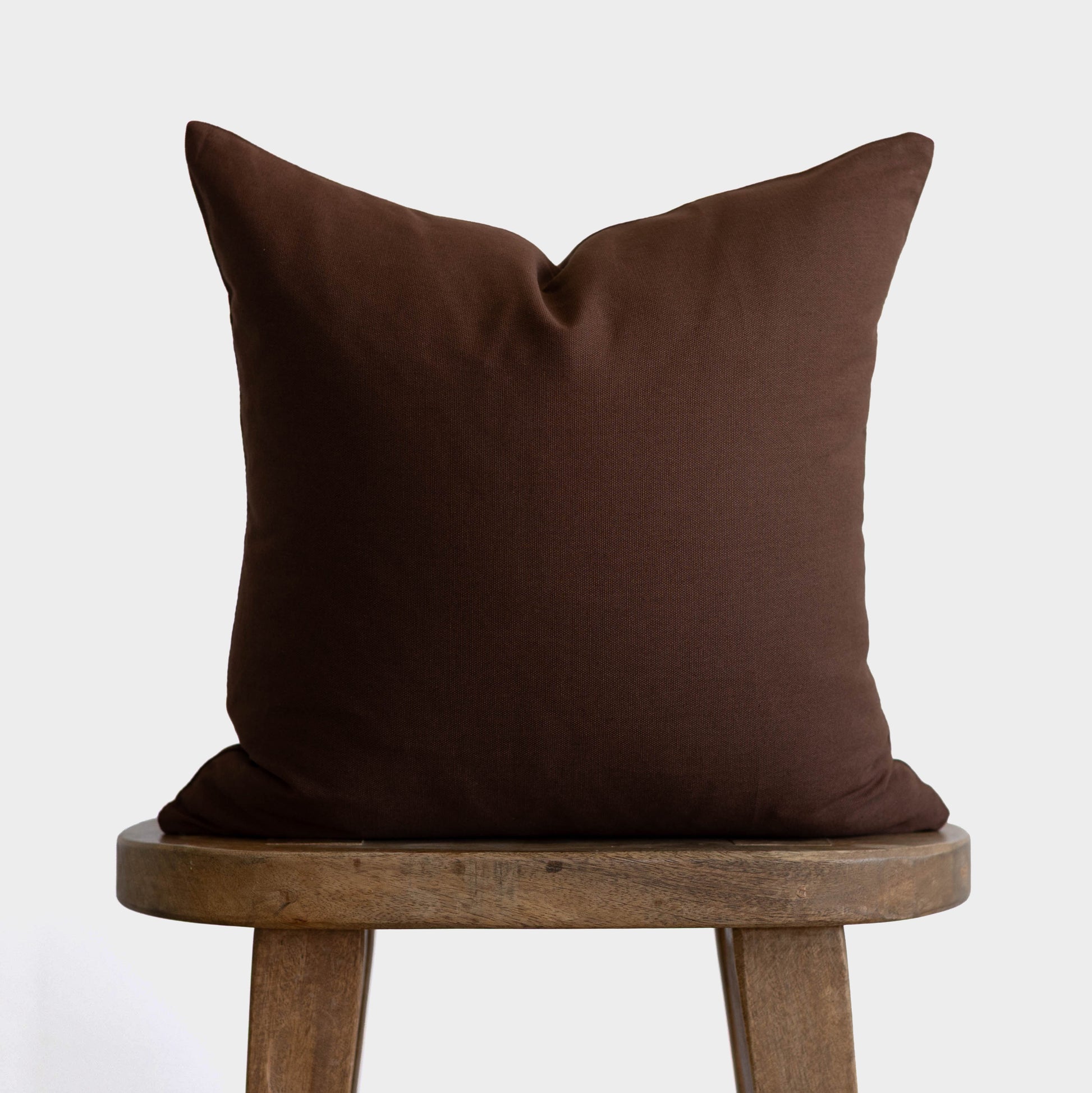 Dark brown linen pillow with envelope closure displayed on wooden stool.