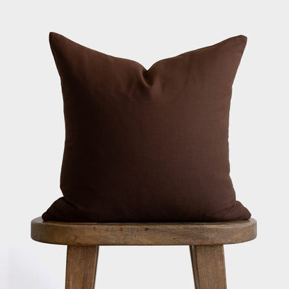 Dark brown linen pillow with envelope closure displayed on wooden stool.