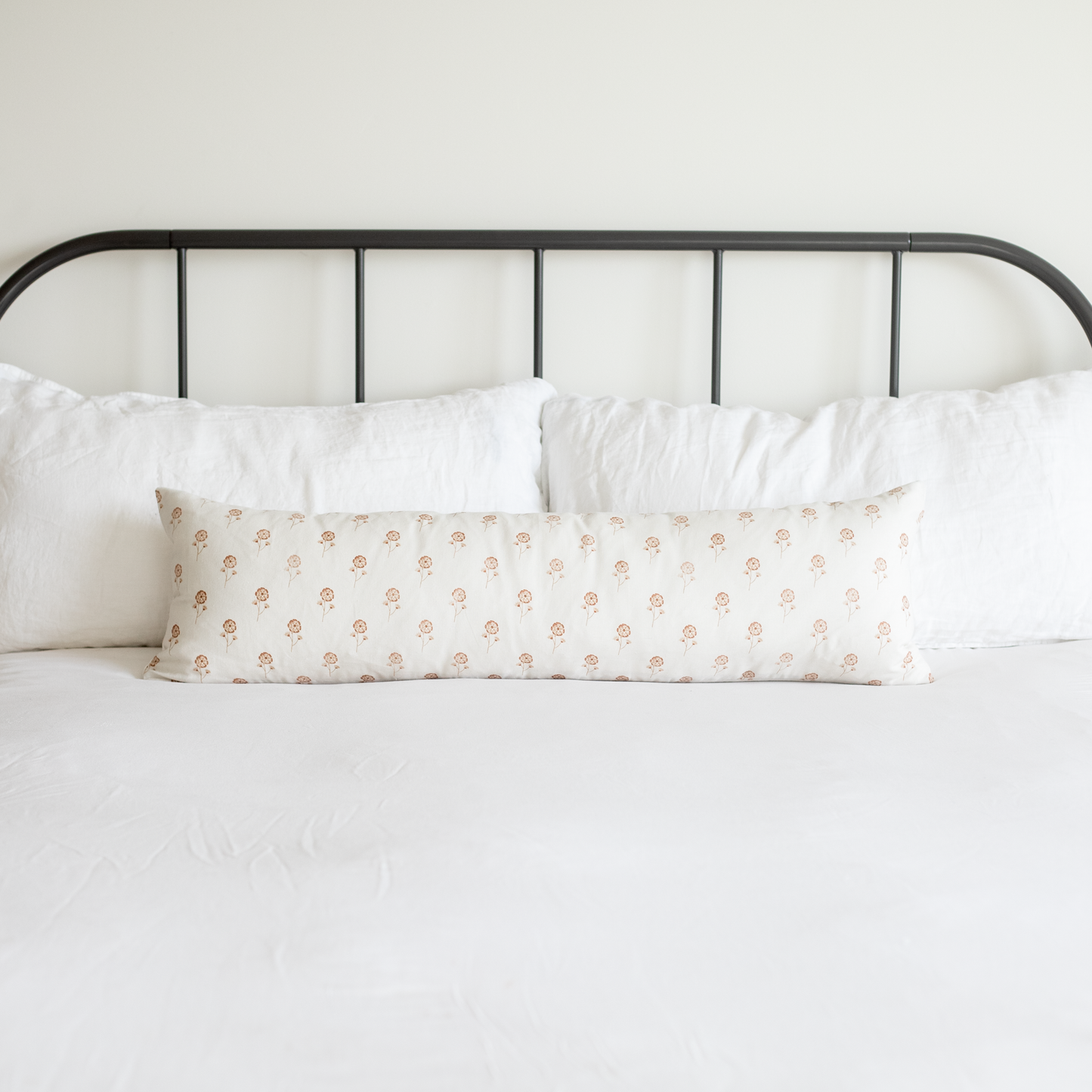 Long lumbar pillow with peach floral pattern on white background, displayed on bed with white pillows and black metal headboa