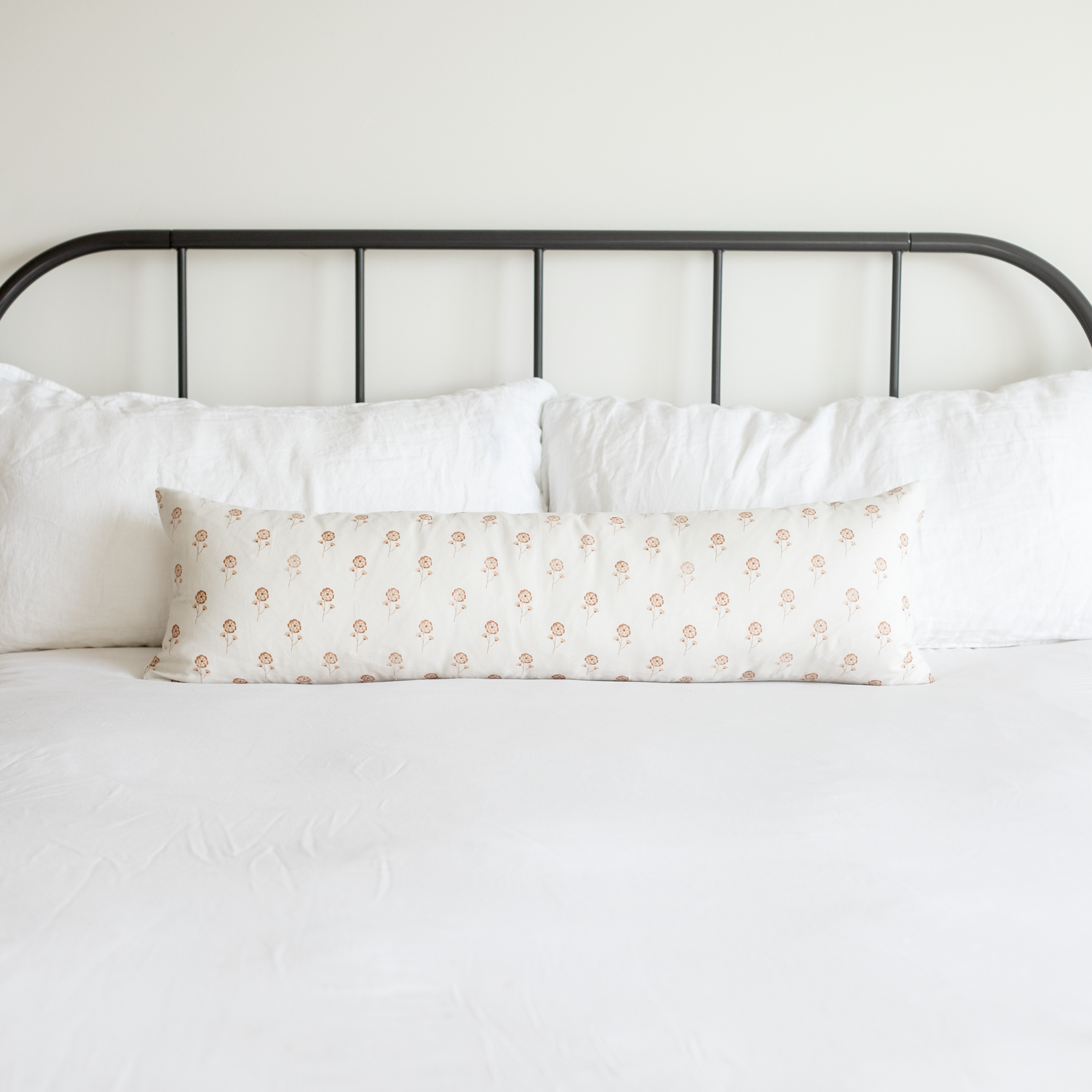 Long lumbar pillow with peach floral pattern on white background, displayed on bed with white pillows and black metal headboa