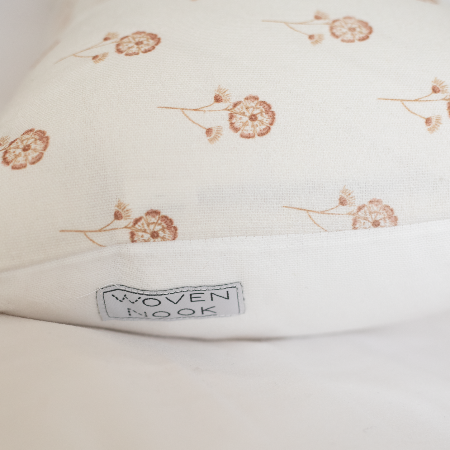 Cream pillow with scattered tan botanical flower motifs and "Woven Nook" label visible on edge.