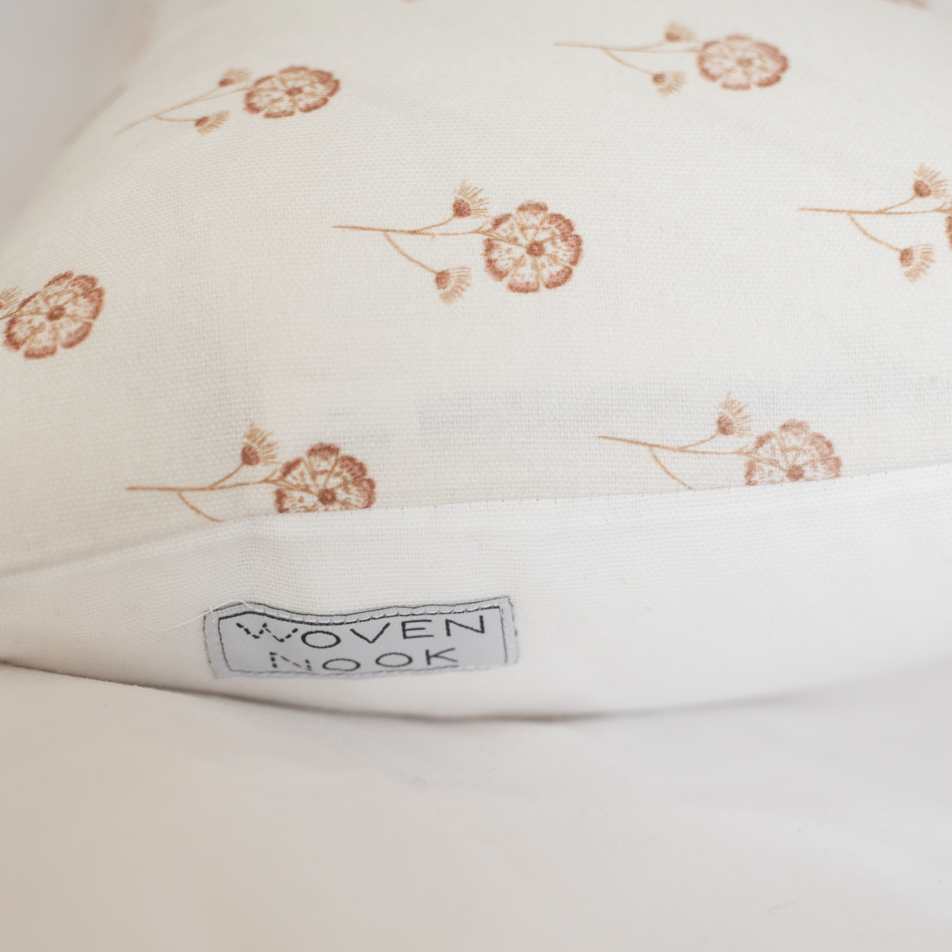 Cream pillow with scattered tan botanical flower motifs and "Woven Nook" label visible on edge.