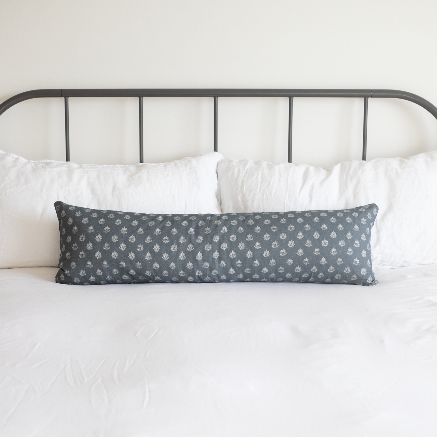 Blue-gray lumbar pillow with repeating white geometric pattern, displayed on bed with white pillows and metal headboard.