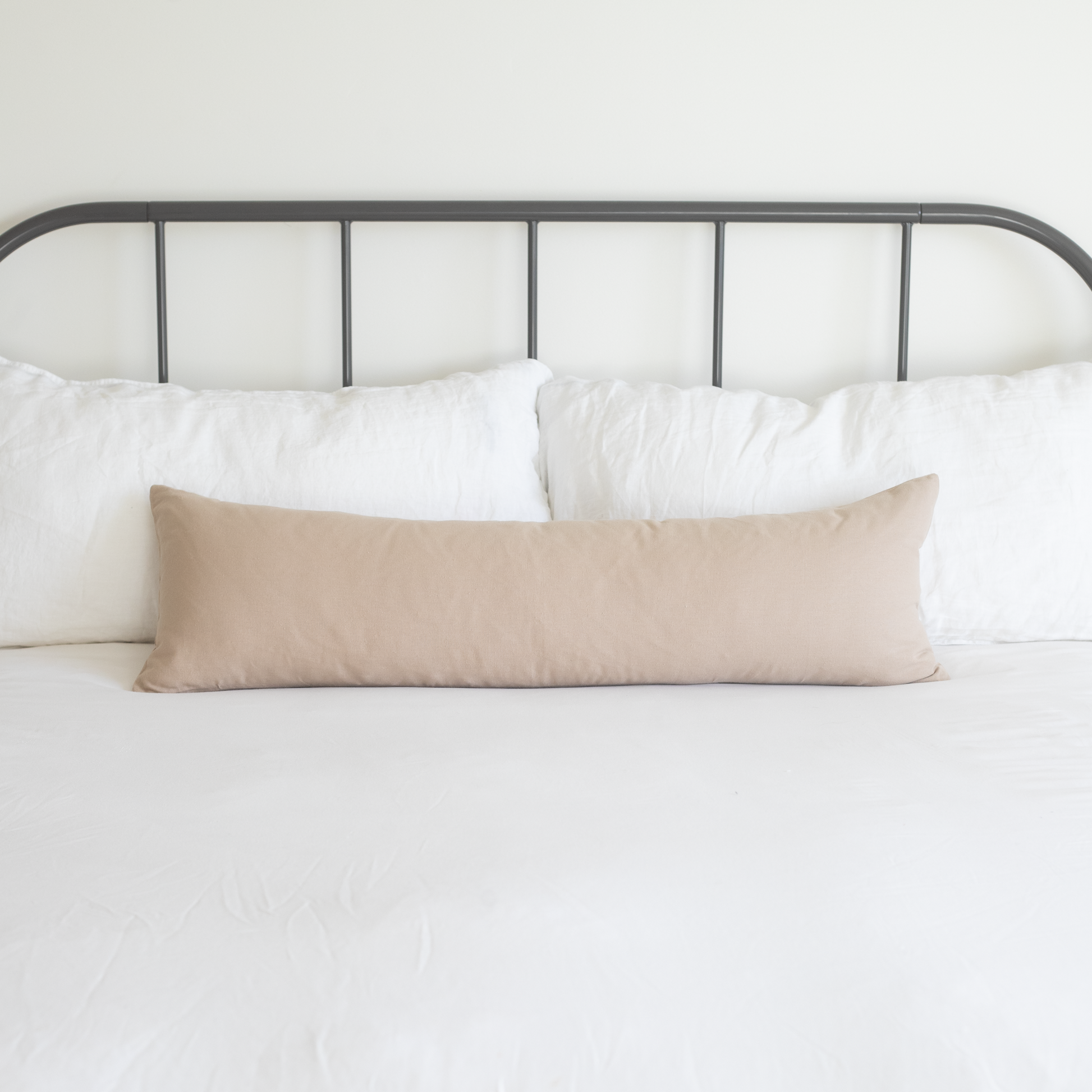 Tan lumbar pillow displayed on a bed with white pillows and metal headboard.