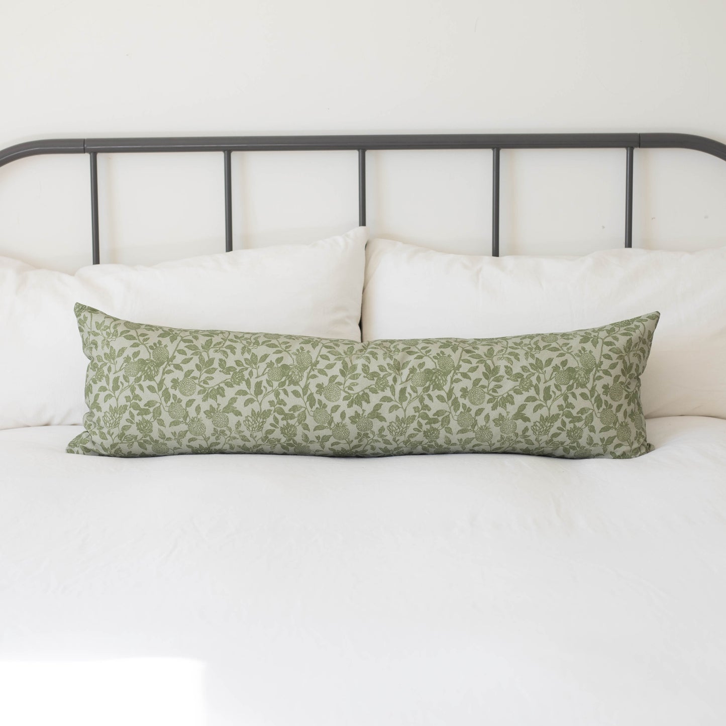 Long rectangular pillow with green botanical leaf pattern on white bedding against metal headboard.