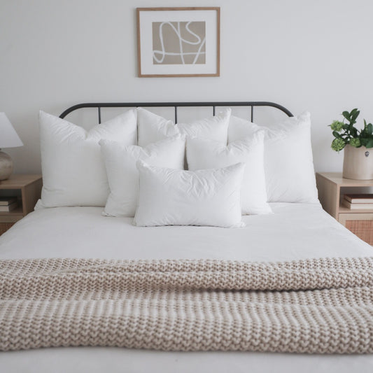 Queen bed with white pillows, white fitted sheet, and beige woven throw blanket on metal frame headboard.