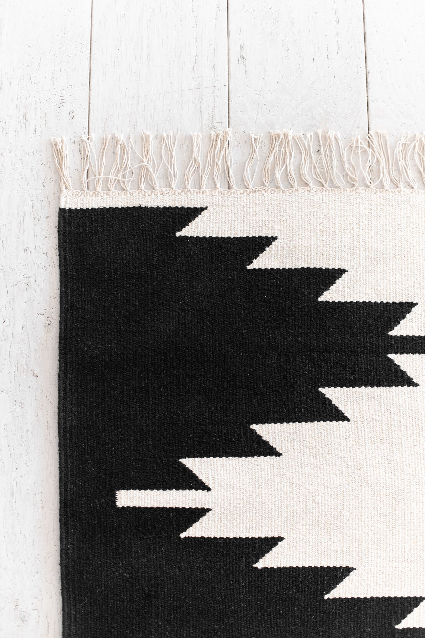 Flat-woven rug with cream and black geometric zigzag pattern, cream fringe trim, on white wooden floor.
