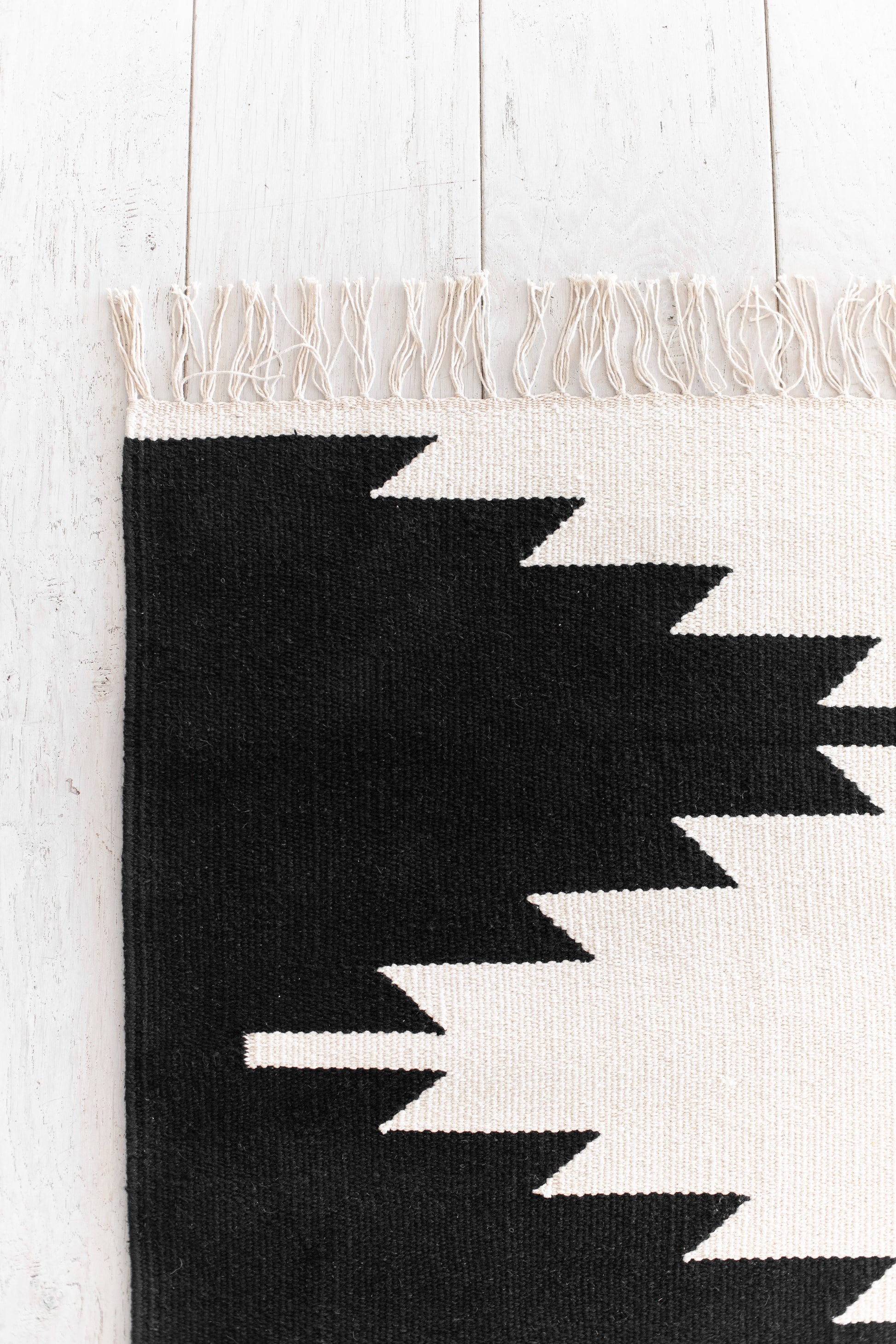 Flat-woven rug with cream and black geometric zigzag pattern, cream fringe trim, on white wooden floor.