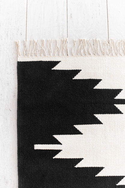 Flat-woven rug with cream and black geometric zigzag pattern, cream fringe trim, on white wooden floor.