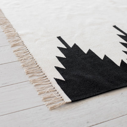 Cream and black geometric patterned area rug with fringe trim on white wooden floor.
