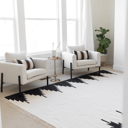 Cream and black geometric patterned area rug displayed in a bright living room with two cream armchairs and a side table.