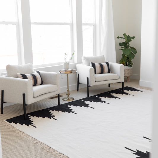 Cream and black geometric patterned area rug displayed in a bright living room with two cream armchairs and a side table.