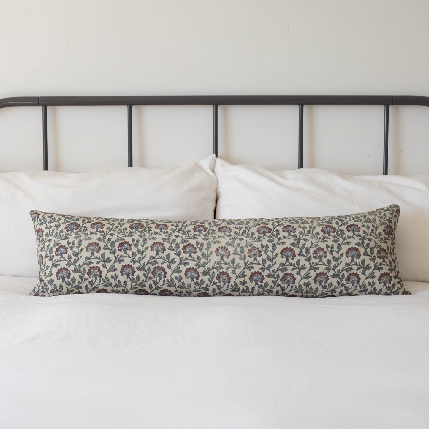 Multicolor floral patterned lumbar pillow with burgundy and blue flowers on cream background, displayed on bed with metal hea