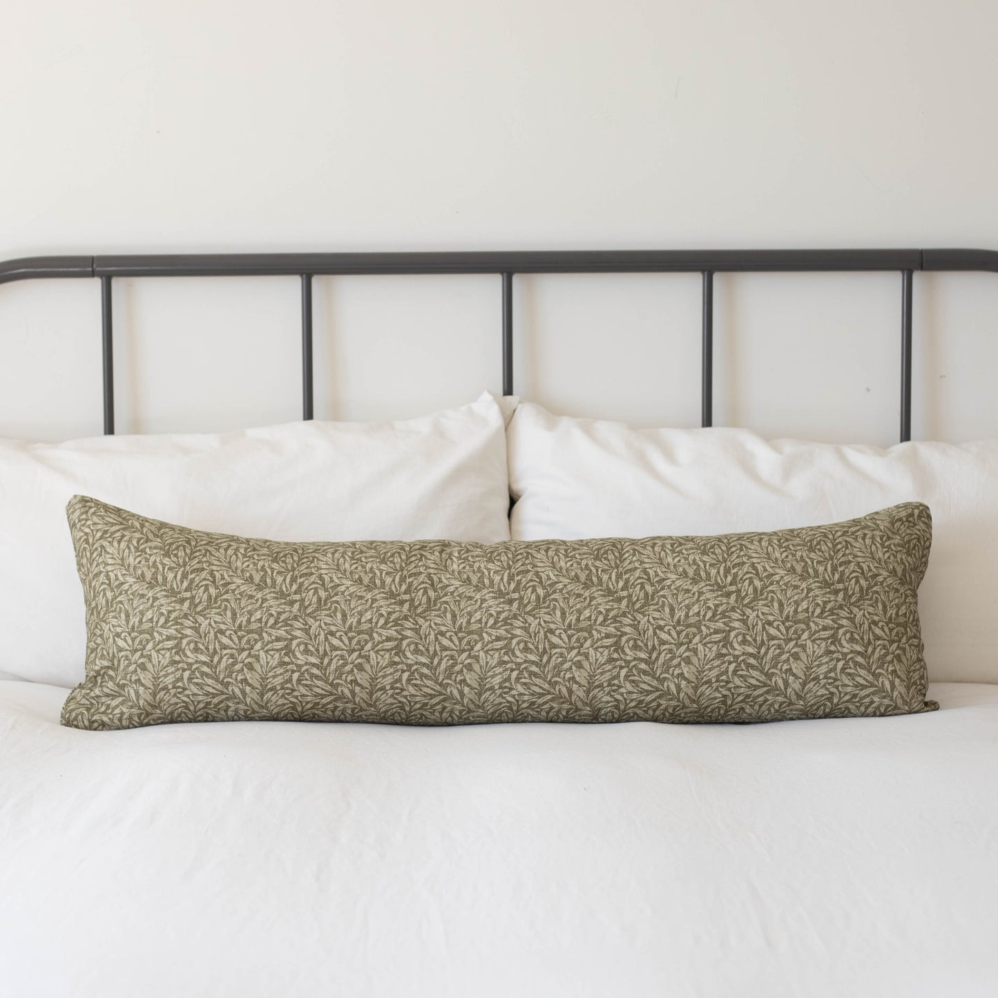 Long rectangular lumbar pillow with green and cream damask pattern on white bedding with black metal headboard.