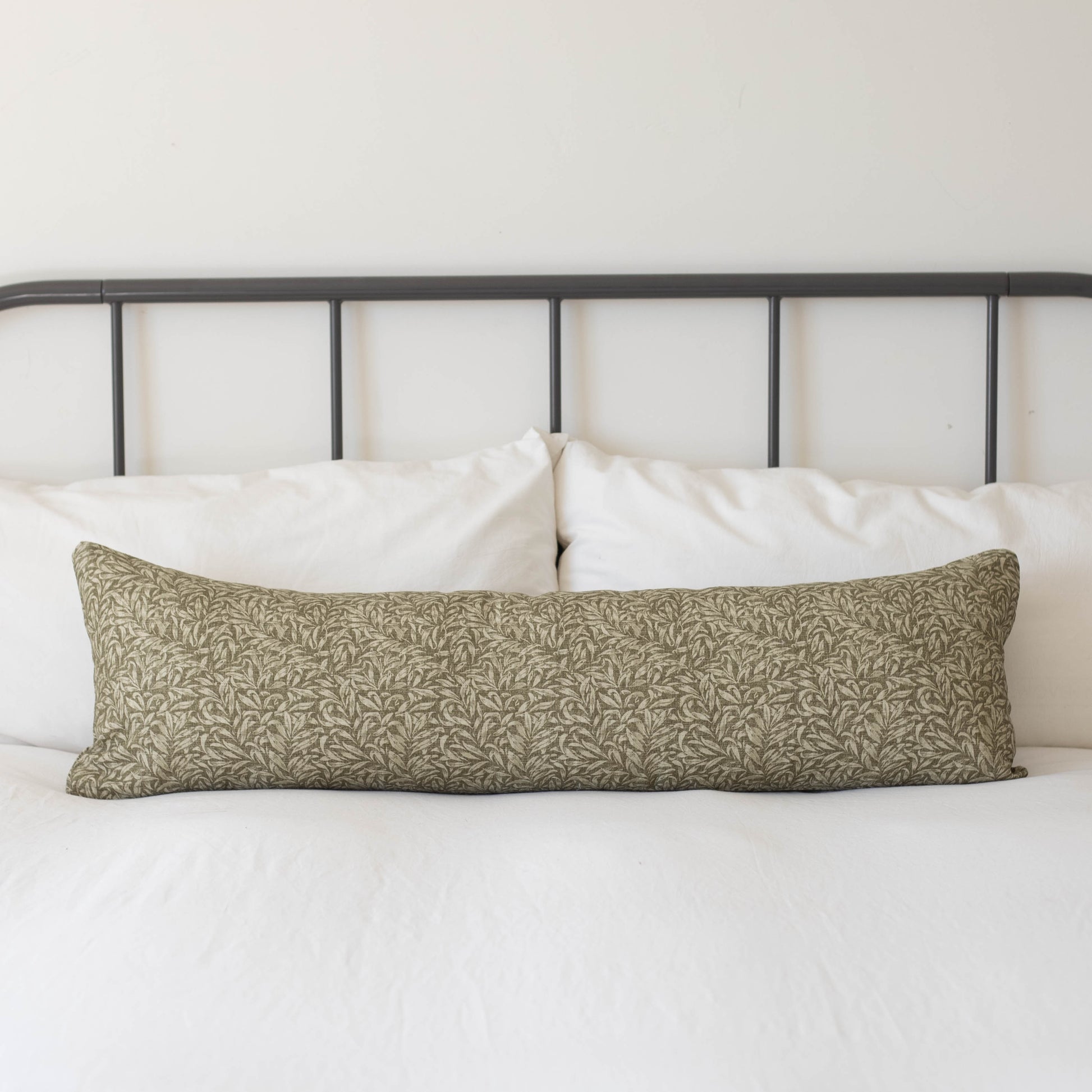 Long rectangular lumbar pillow with green and cream damask pattern on white bedding with black metal headboard.