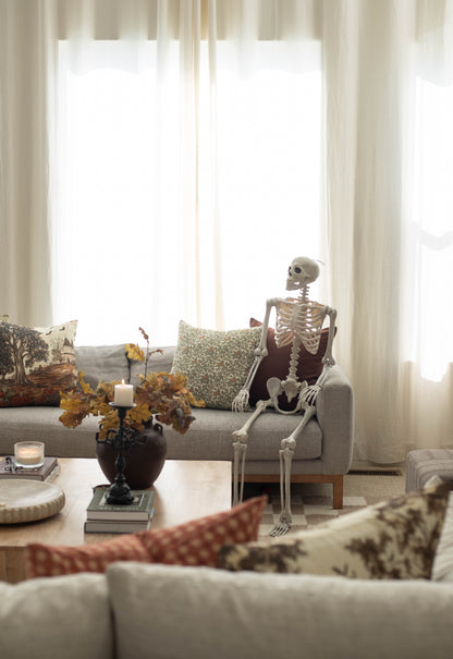 Cream linen pillow with dark autumn foliage and bird motif displayed on gray sofa with decorative skeleton figure.