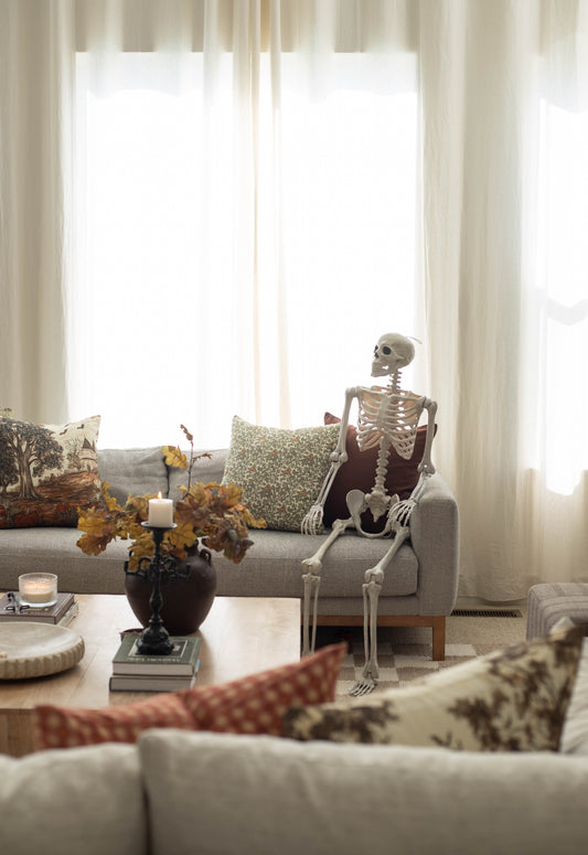 Cream linen pillow with dark autumn foliage and bird motif displayed on gray sofa with decorative skeleton figure.