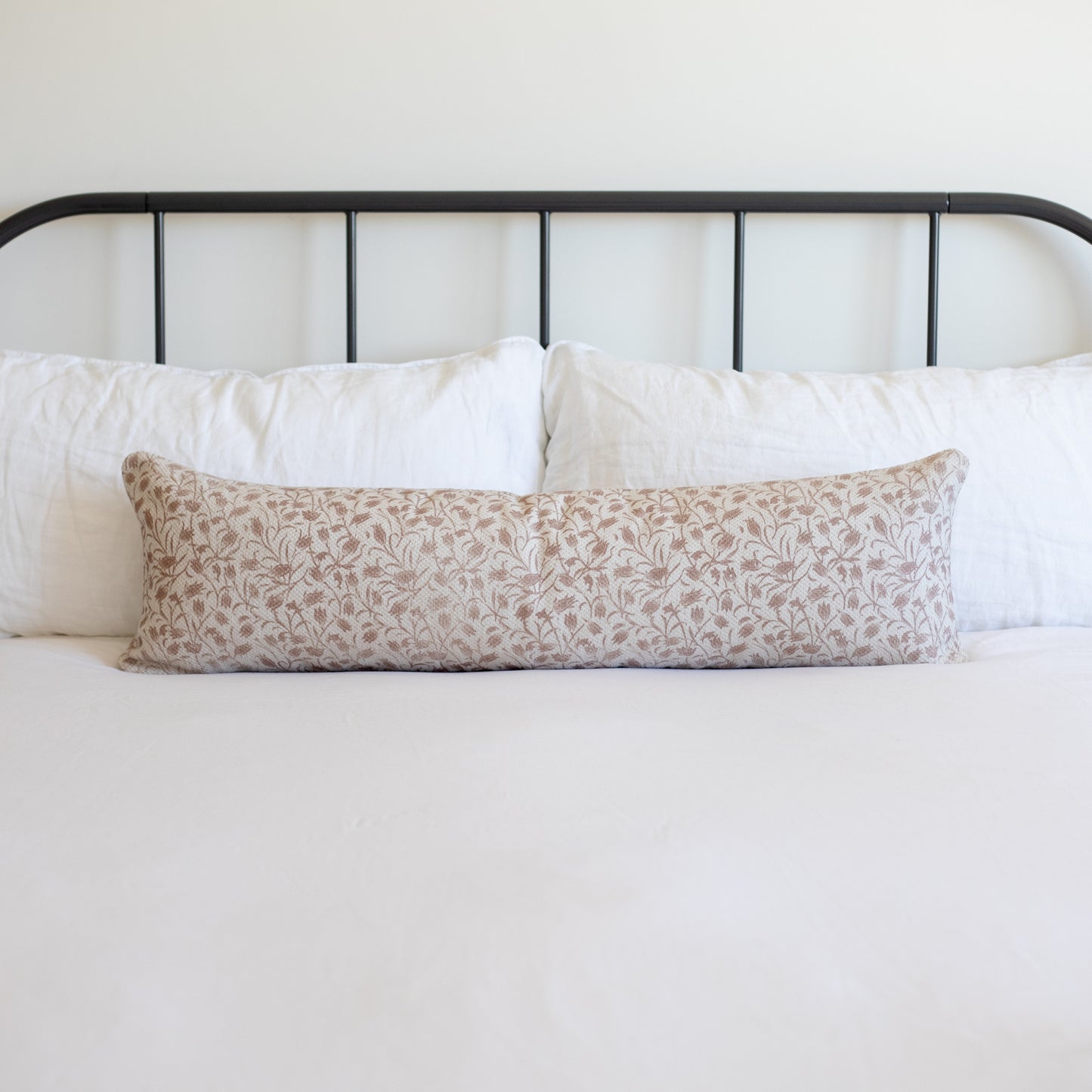 Rectangular throw pillow with tan and white leaf pattern on bed with white pillows and black metal headboard.