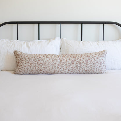 Rectangular throw pillow with tan and white leaf pattern on bed with white pillows and black metal headboard.