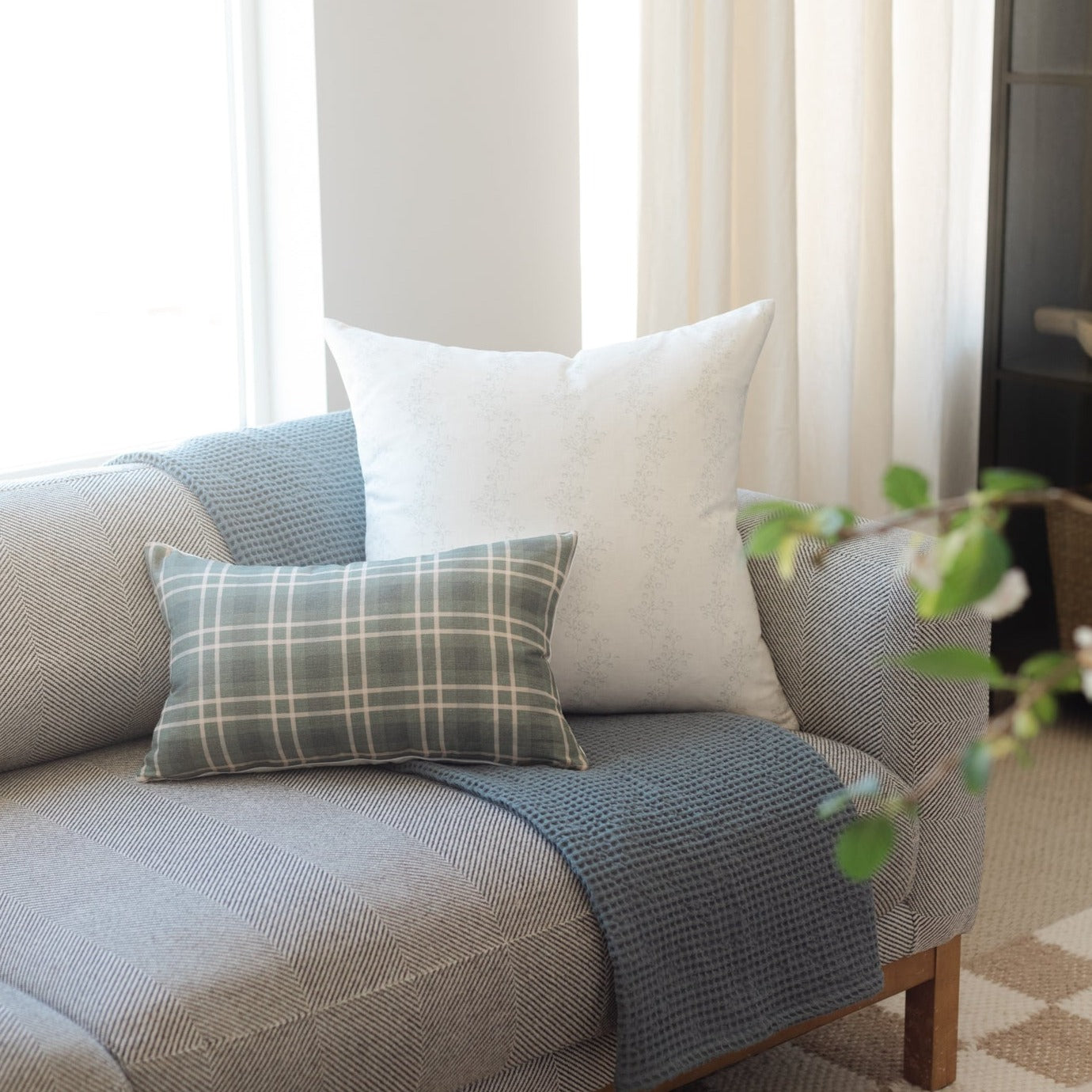 Three decorative pillows arranged on a blue upholstered sofa: a gray plaid rectangular pillow, a cream square pillow, and a t