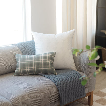 Three decorative pillows arranged on a blue upholstered sofa: a gray plaid rectangular pillow, a cream square pillow, and a t