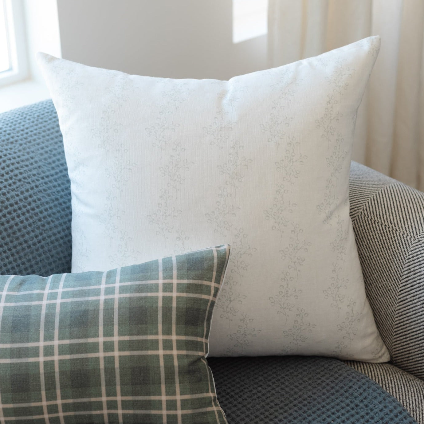 White decorative pillow with subtle leaf pattern, displayed on furniture with plaid and textured pillows.