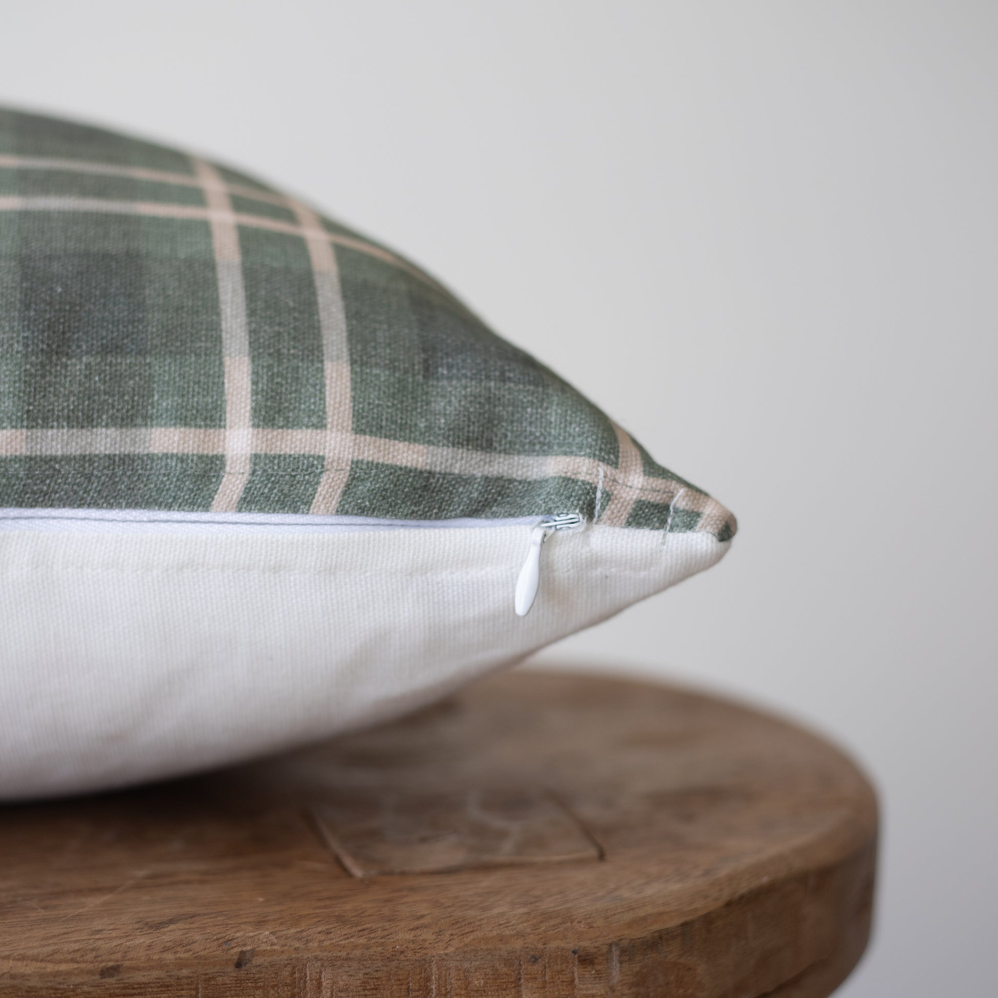 Green and white plaid pillow with white zipper detail resting on wooden round surface.