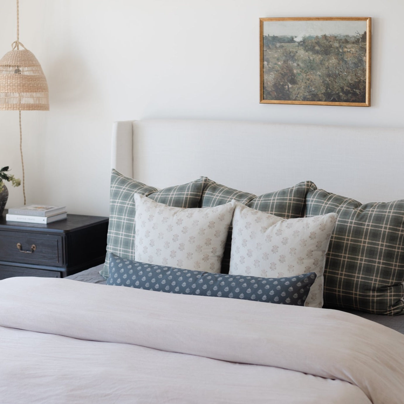 Framed landscape artwork with wood border hanging above a bed with patterned pillows and white bedding.