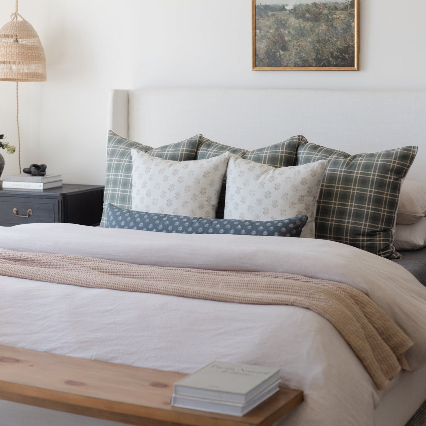 Bed styled with white linens, tan textured throw, and assorted plaid and patterned pillows in neutral tones.