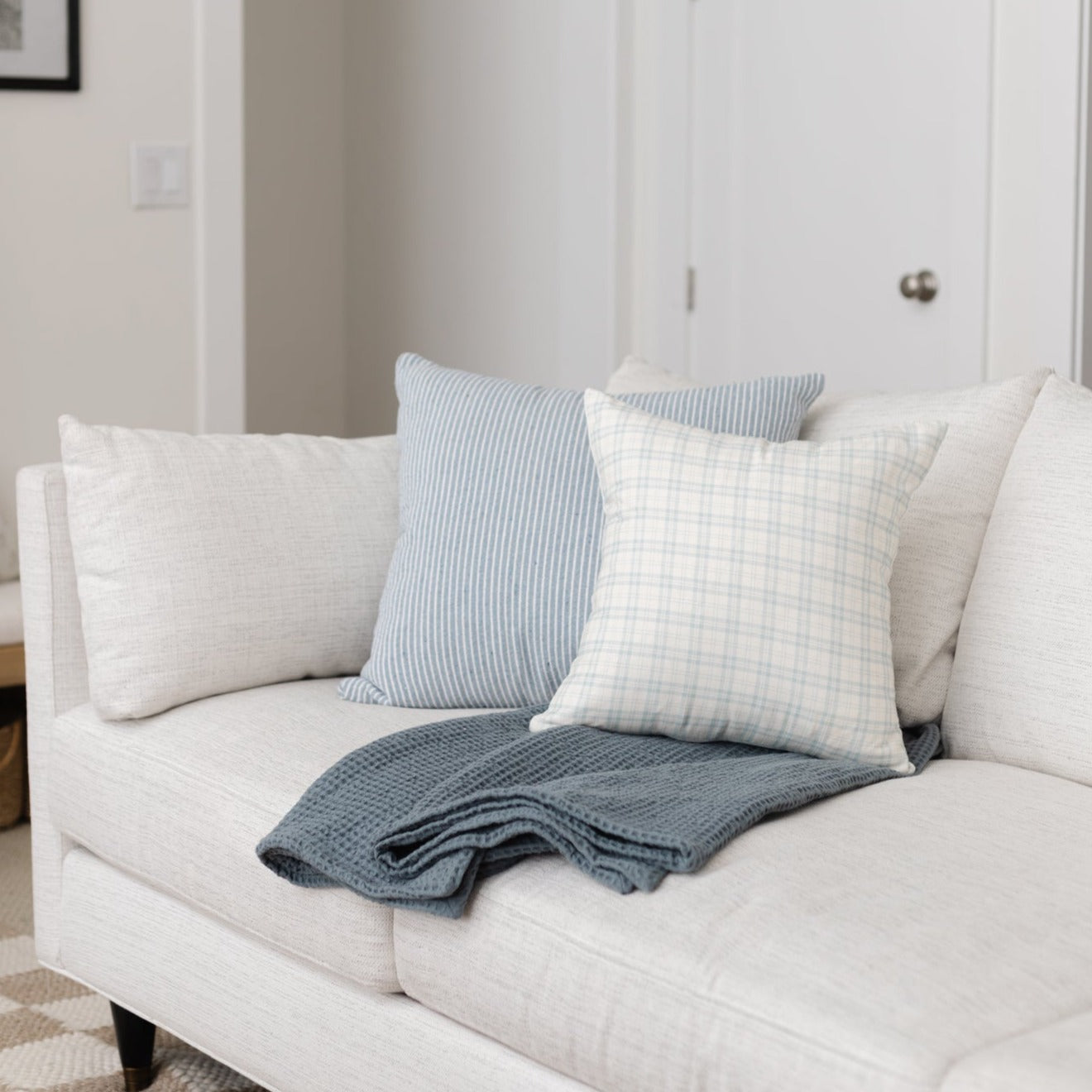 Cream upholstered sofa with blue striped and checkered throw pillows and folded blue knit blanket.
