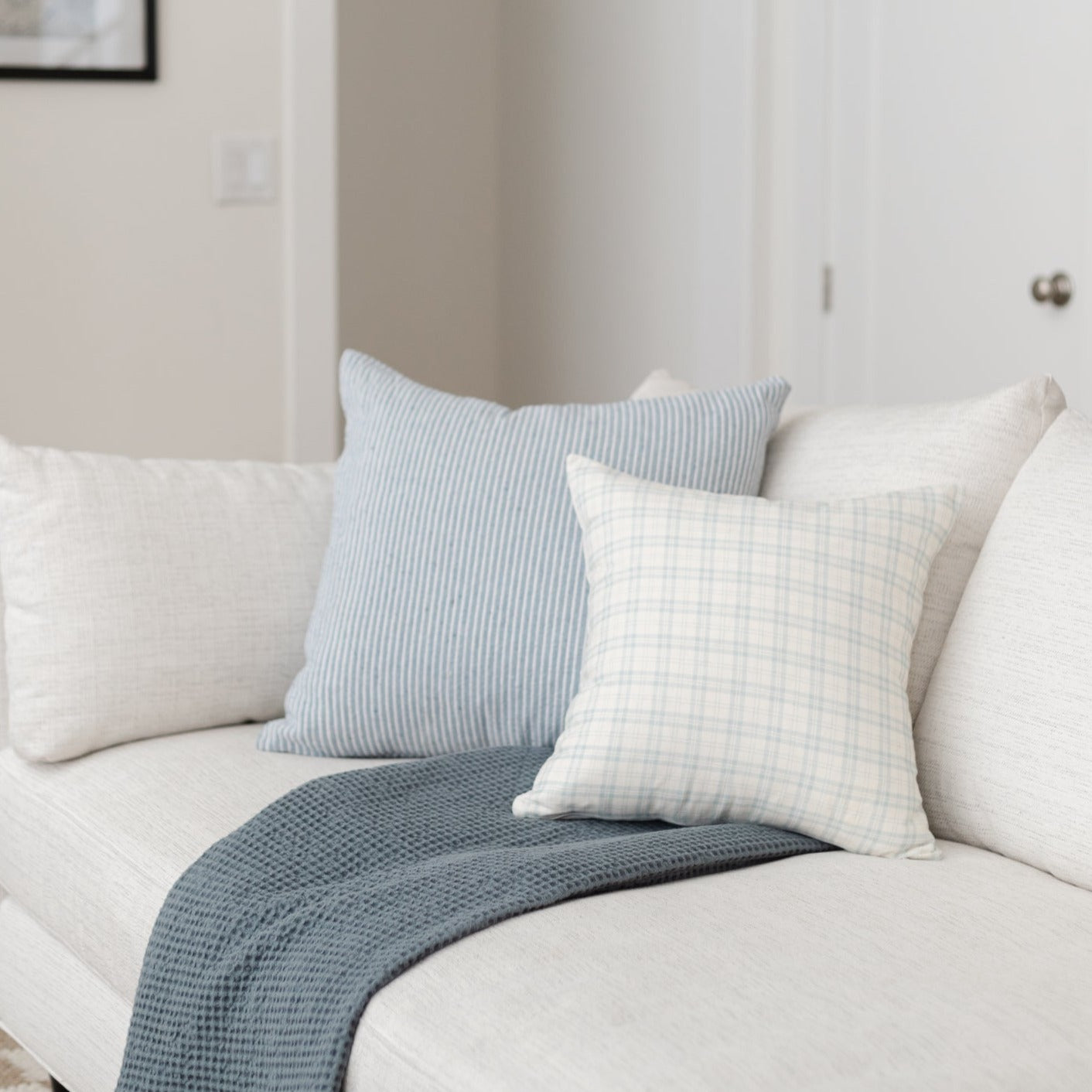 Cream upholstered sofa with blue striped, cream plaid, and cream textured throw pillows and folded blue knit blanket.