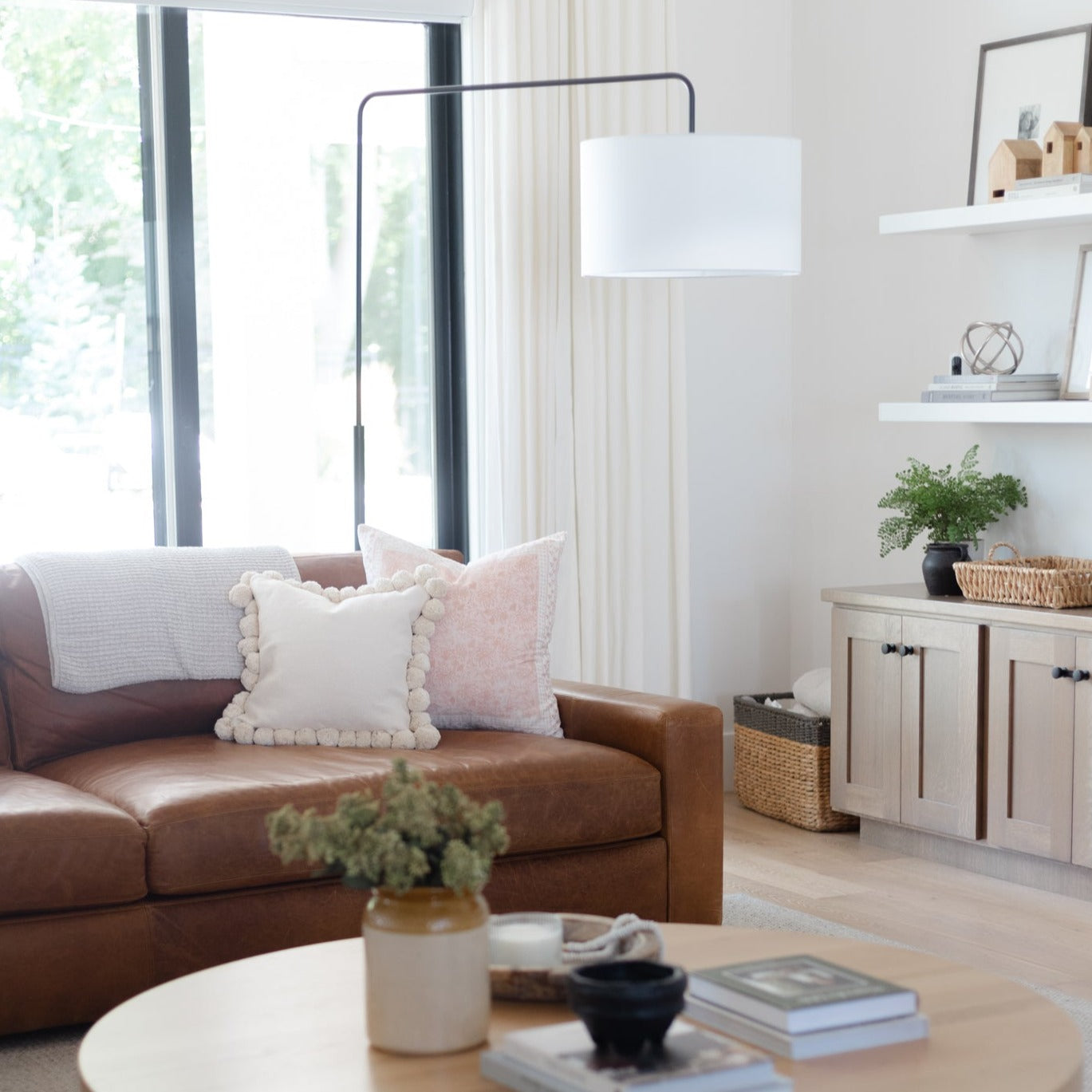 Arc floor lamp with curved black frame and white cylindrical shade suspended over brown leather sofa.