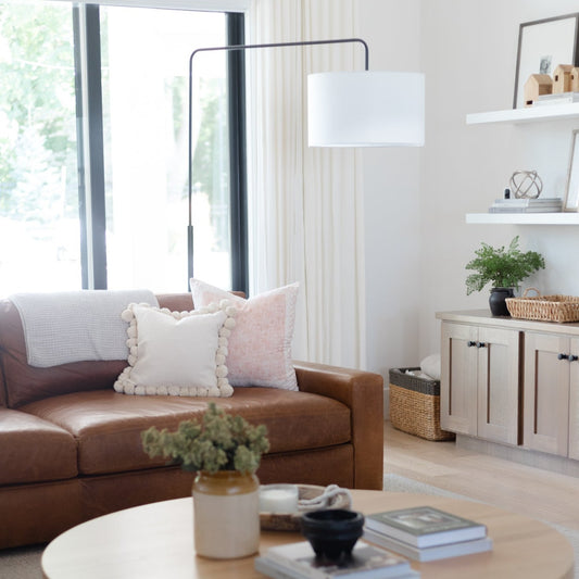 Arc floor lamp with curved black frame and white cylindrical shade suspended over brown leather sofa.
