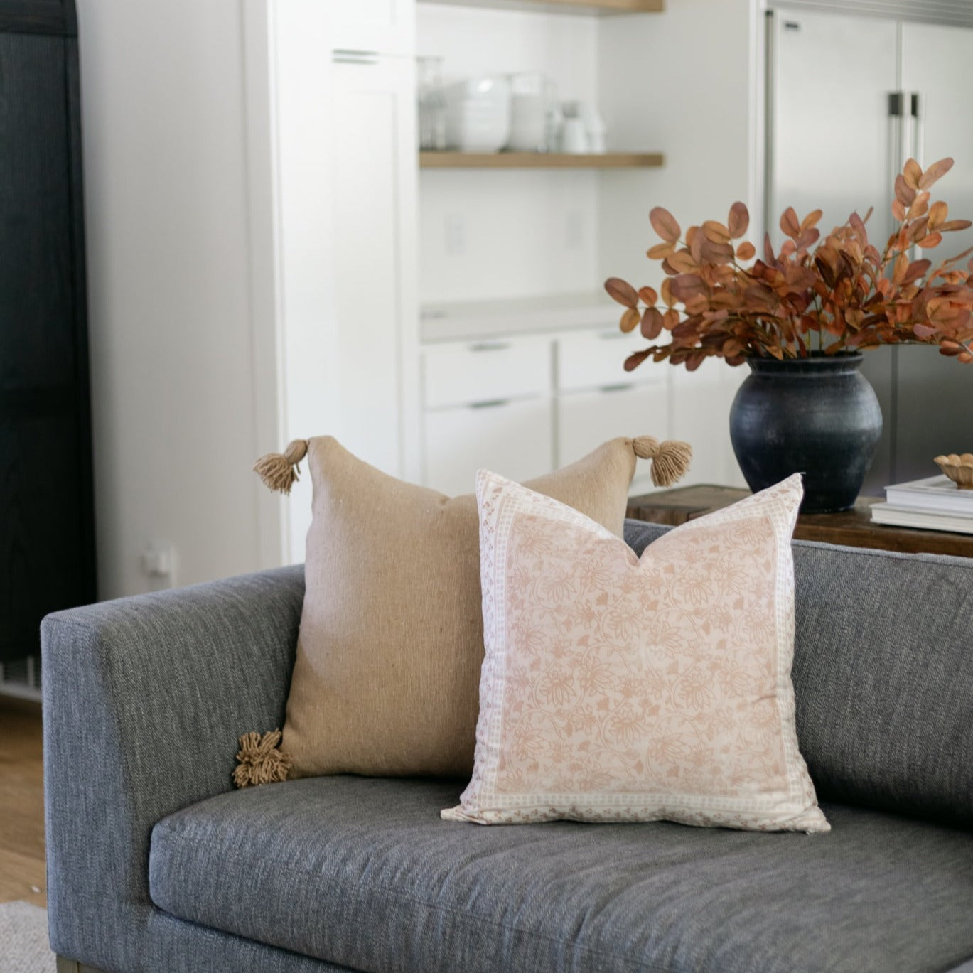 Two throw pillows with tassel details on gray sofa, one tan solid and one cream patterned.