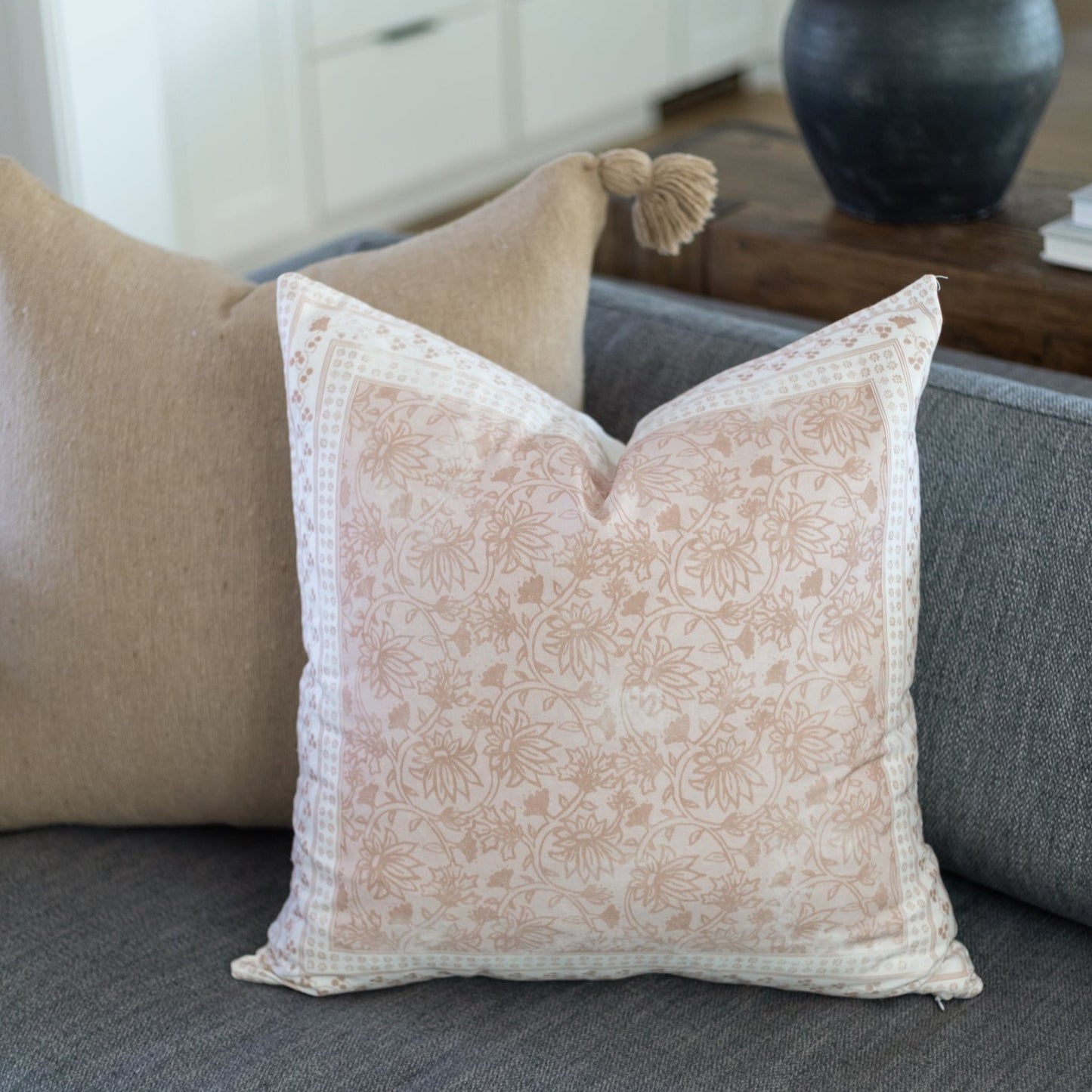 Cream and tan damask patterned pillow with decorative tassels on a gray sofa.