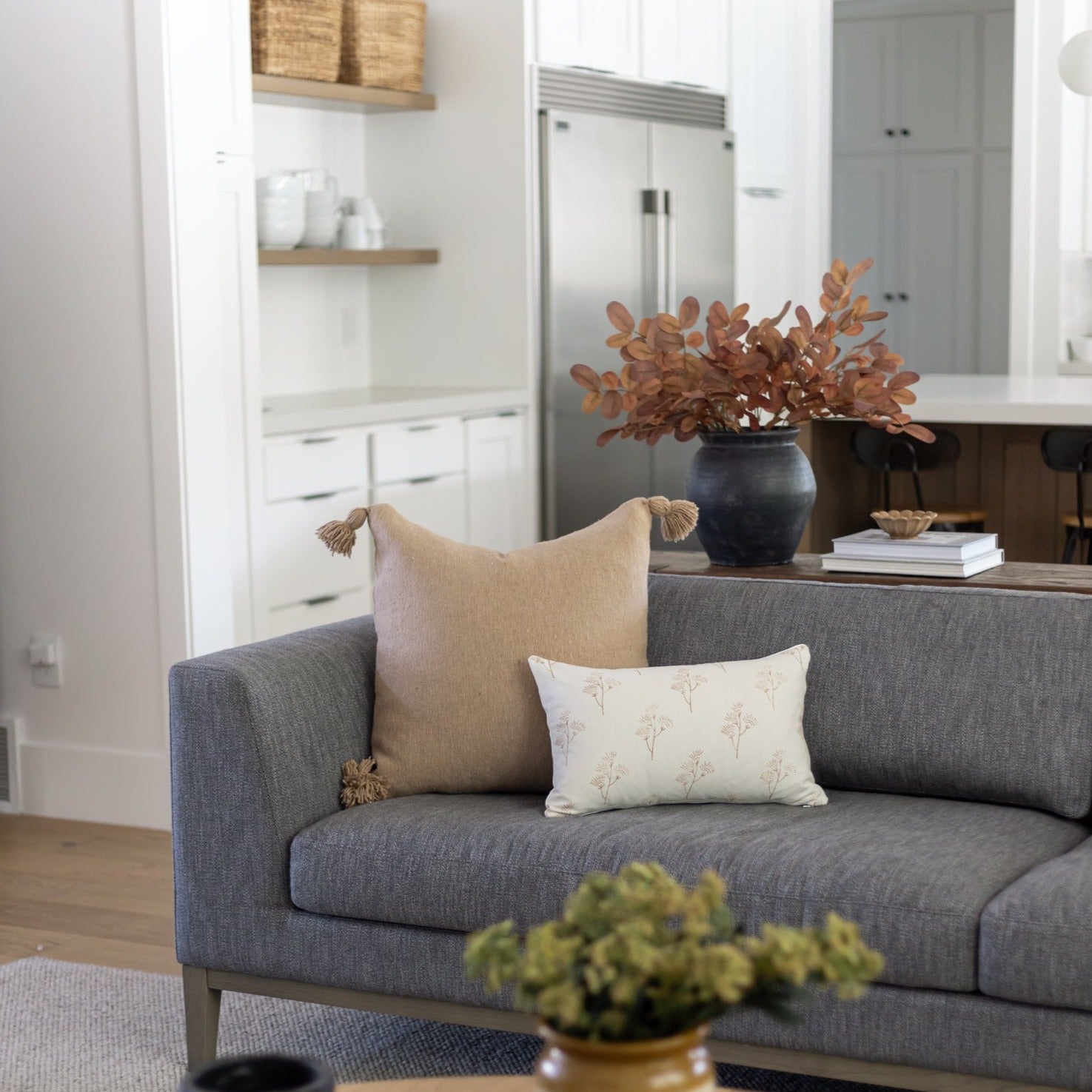 Cream lumbar pillow with tassel corners displayed on gray sofa with thistle pattern.