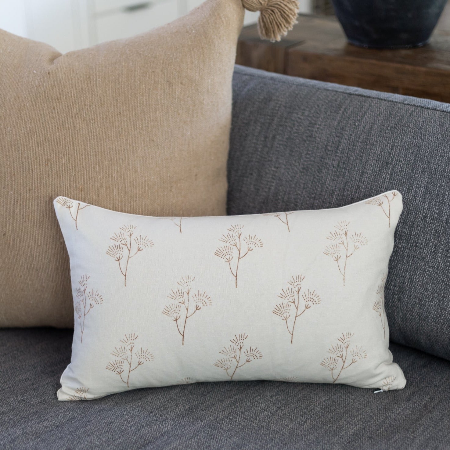 Cream lumbar pillow with tan thistle plant print, displayed on gray sofa with beige pillow.
