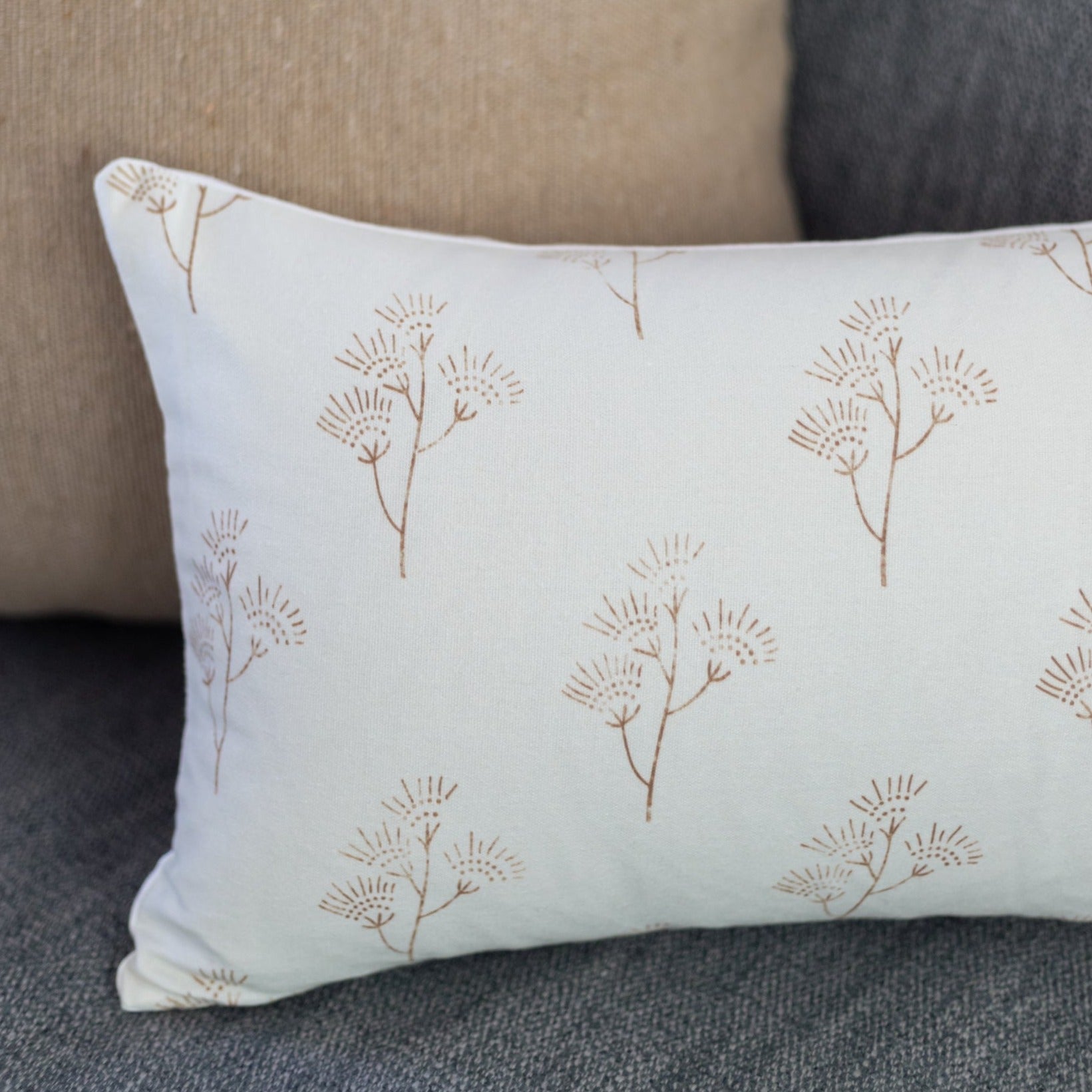 Cream lumbar pillow with tan thistle plant pattern, displayed on gray upholstered furniture.