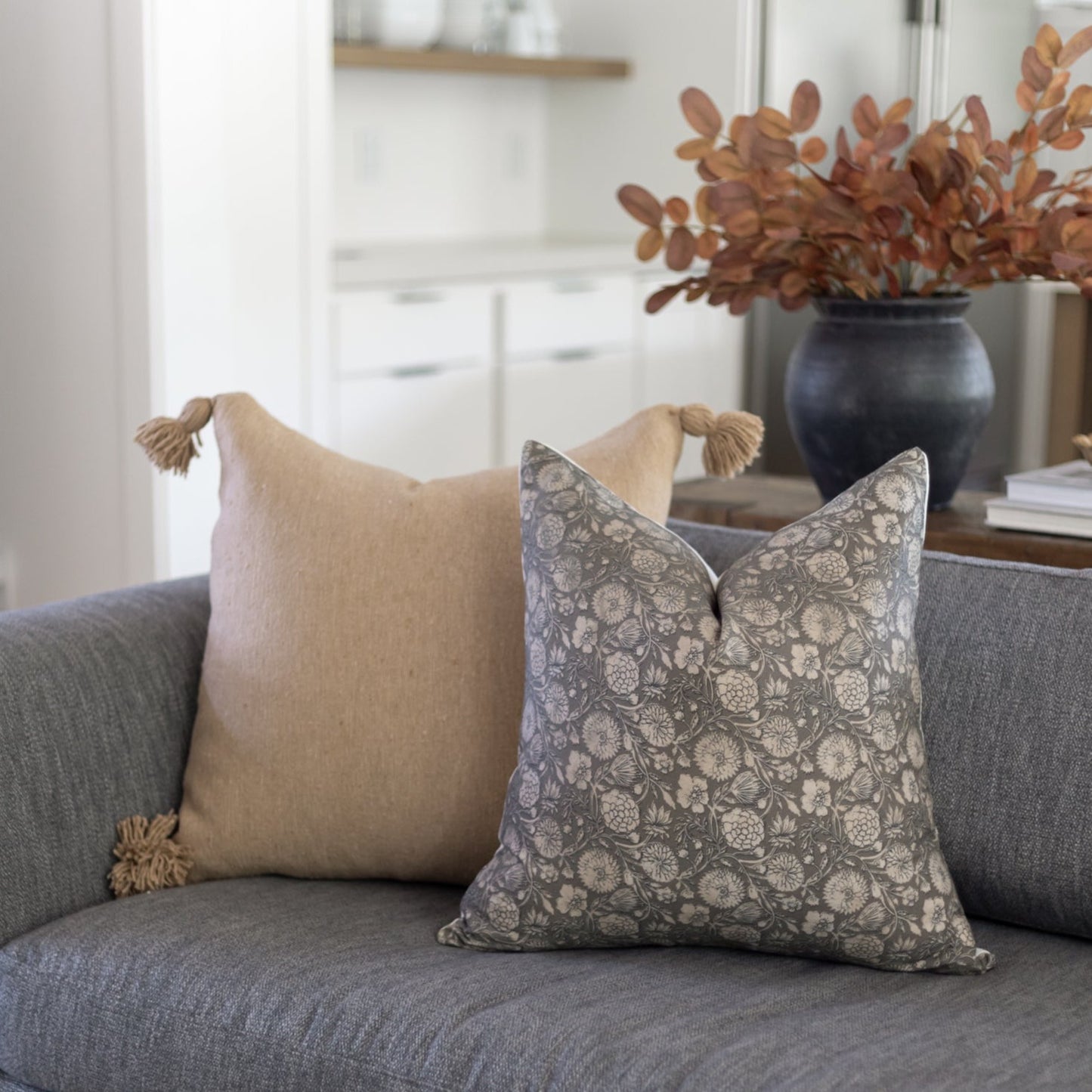 Two decorative pillows on gray sofa: beige linen with tassels and gray floral patterned pillow.