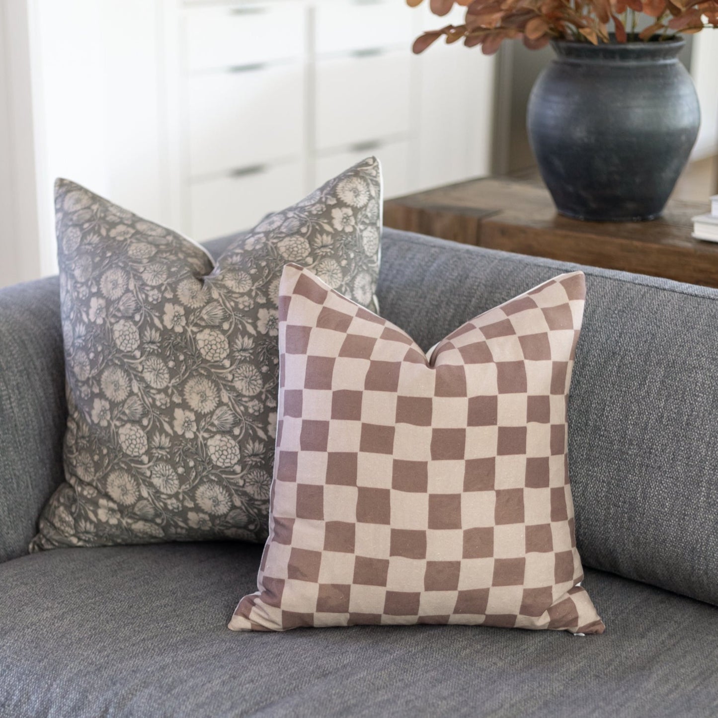Two decorative pillows on a gray sofa: one with gray floral damask pattern, one with mauve and cream checkerboard design.