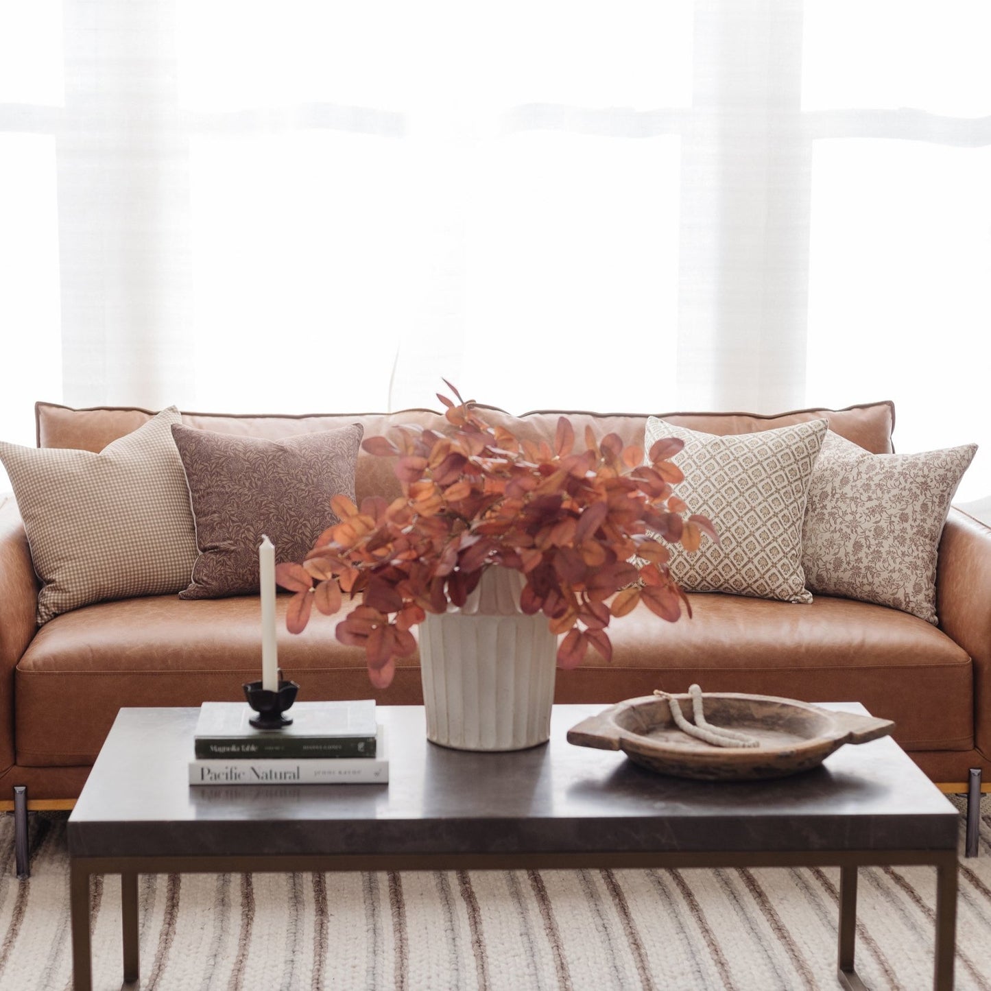 Tan leather sofa with patterned pillows, positioned behind a dark coffee table with books, candle, and decorative bowl.