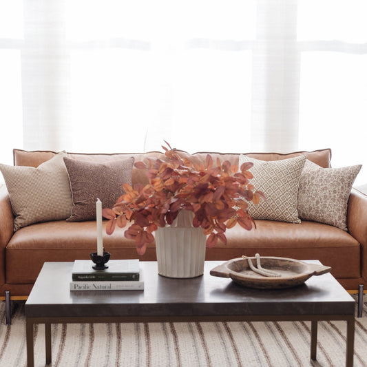 Tan leather sofa with patterned pillows, positioned behind a dark coffee table with books, candle, and decorative bowl.