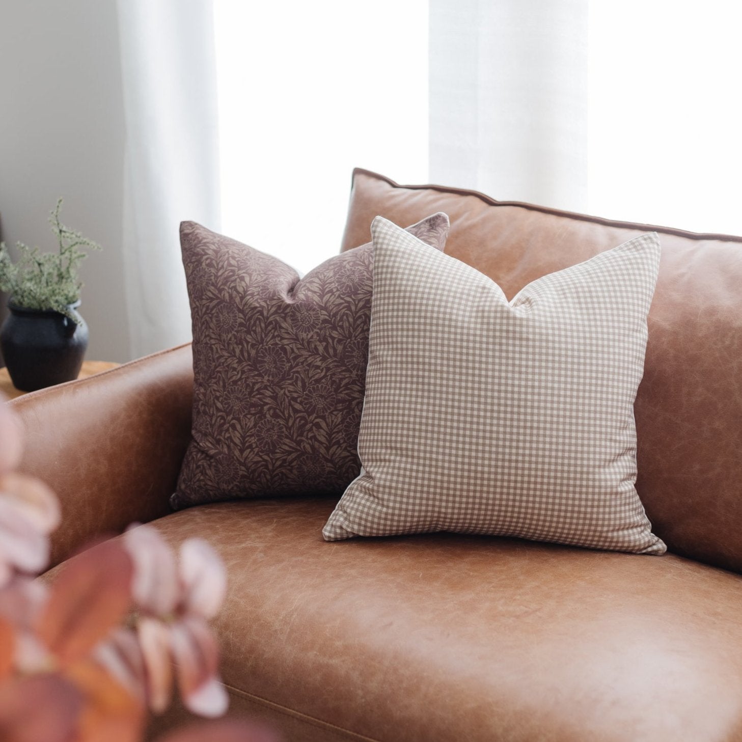 Two decorative pillows with damask and gingham patterns displayed on a cognac leather sofa.