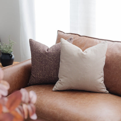 Two decorative pillows with damask and gingham patterns displayed on a tan leather armchair.