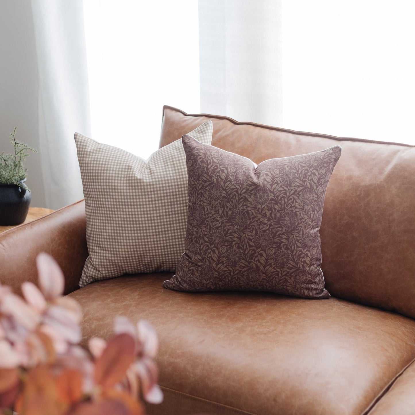 Four throw pillows in taupe gingham and floral damask patterns displayed on a cognac leather sofa.