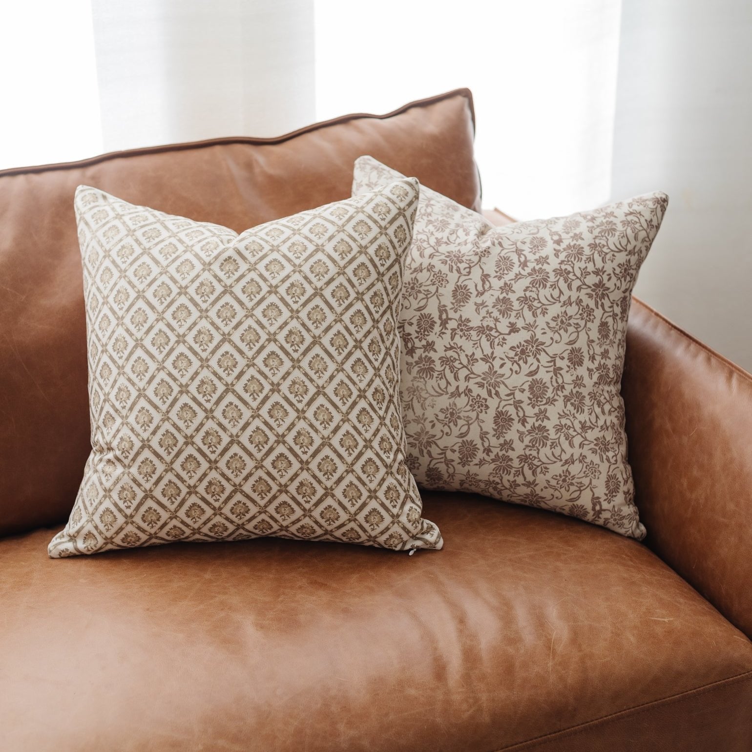 Two patterned throw pillows with geometric and floral designs in beige and tan tones displayed on a brown leather sofa.
