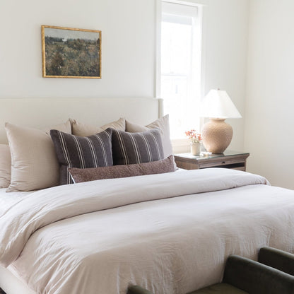 Striped lumbar pillow in charcoal and cream displayed on a bed with neutral bedding and coordinating pillows.