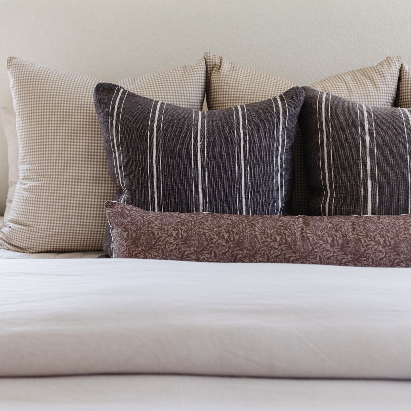 Four lumbar pillows arranged on a bed: two with tan gingham pattern, two with charcoal vertical stripes, and one with brown d
