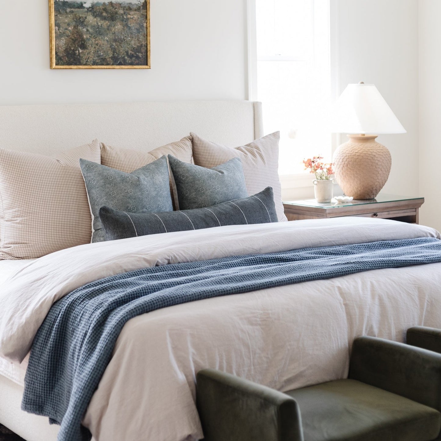 Framed landscape artwork with moss and forest tones displayed above a bedroom bed with blue striped runner and decorative pil